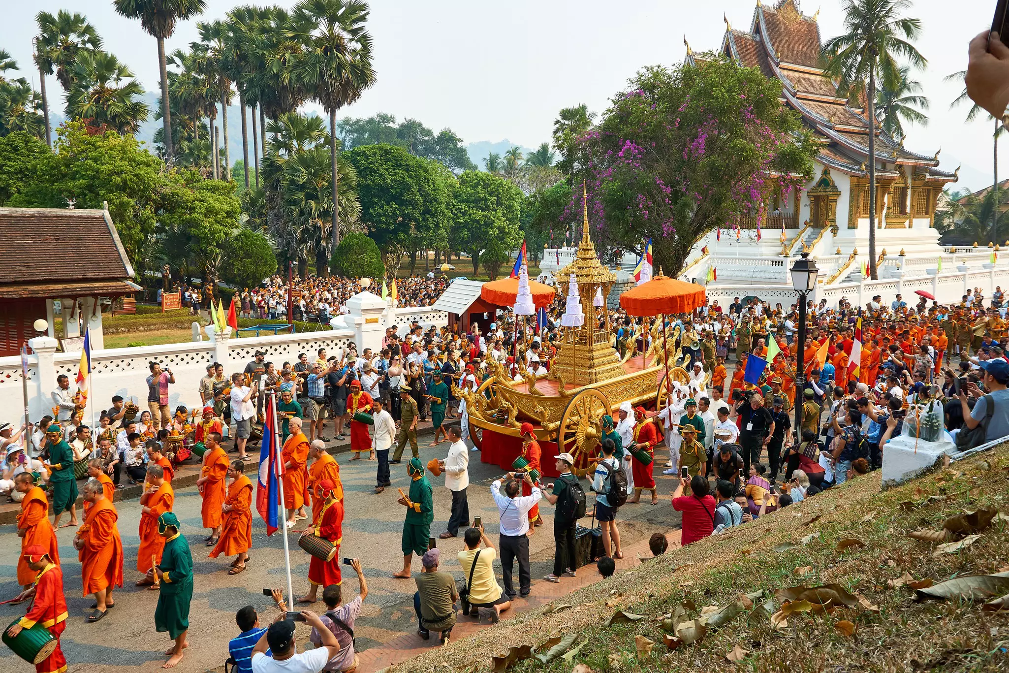 A parade of people in green, orange and red robes surround an ornate red and gold float as it proceeds down a street lined with spectators