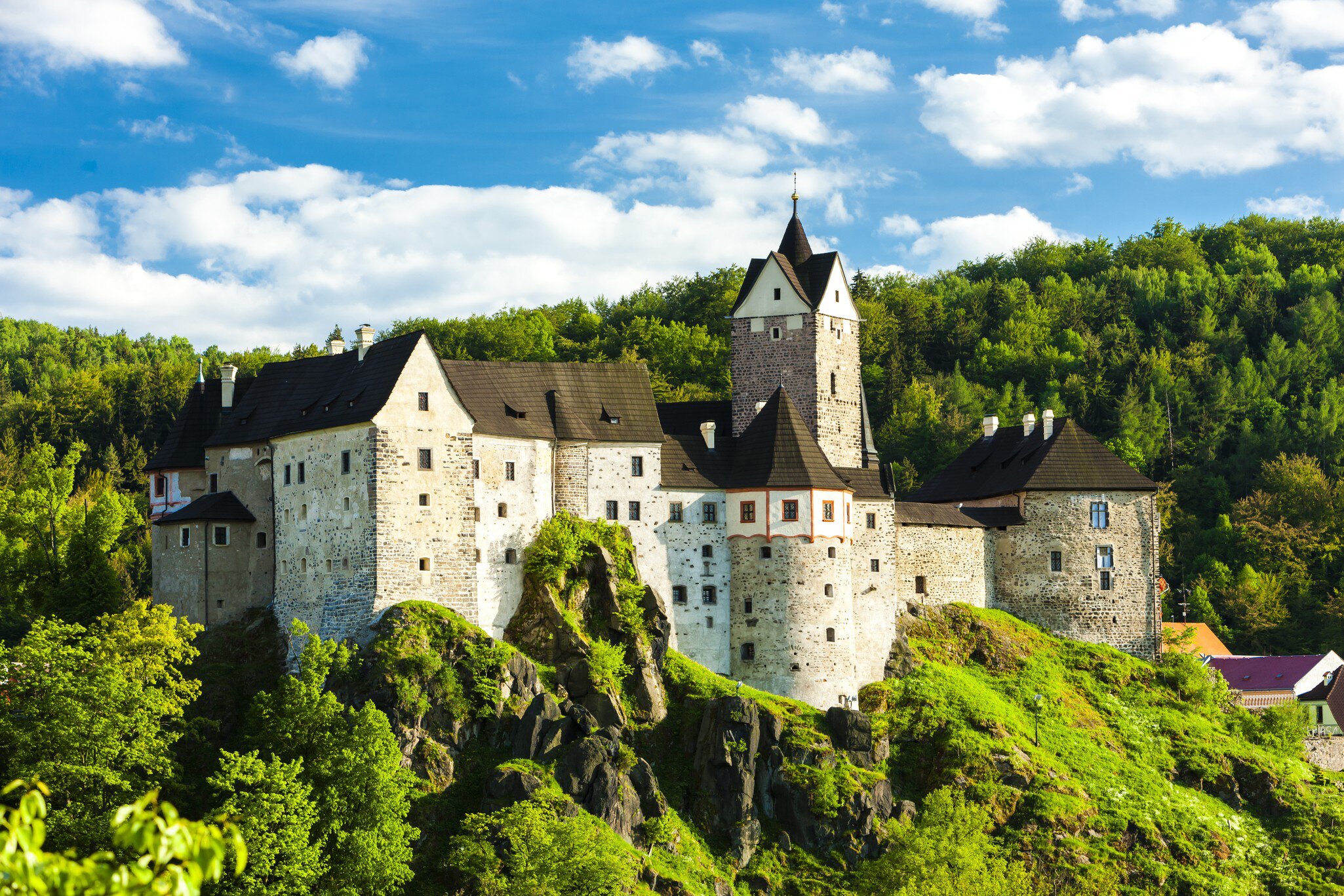 Wide exterior of Loket Castle, Karlovy Vary, Czech Republic. Trees surrounding and bright blue sky.