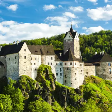 Wide exterior of Loket Castle, Karlovy Vary, Czech Republic. Trees surrounding and bright blue sky.