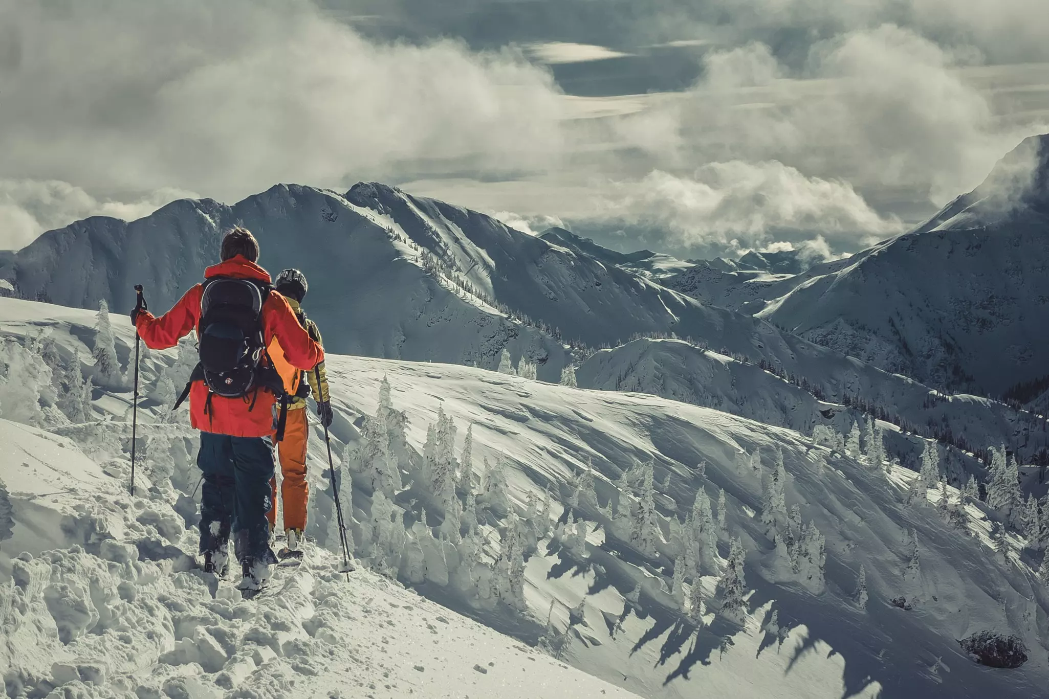 Two backcountry skiers on top of deep snow overlooking mountains on an overcast day.
