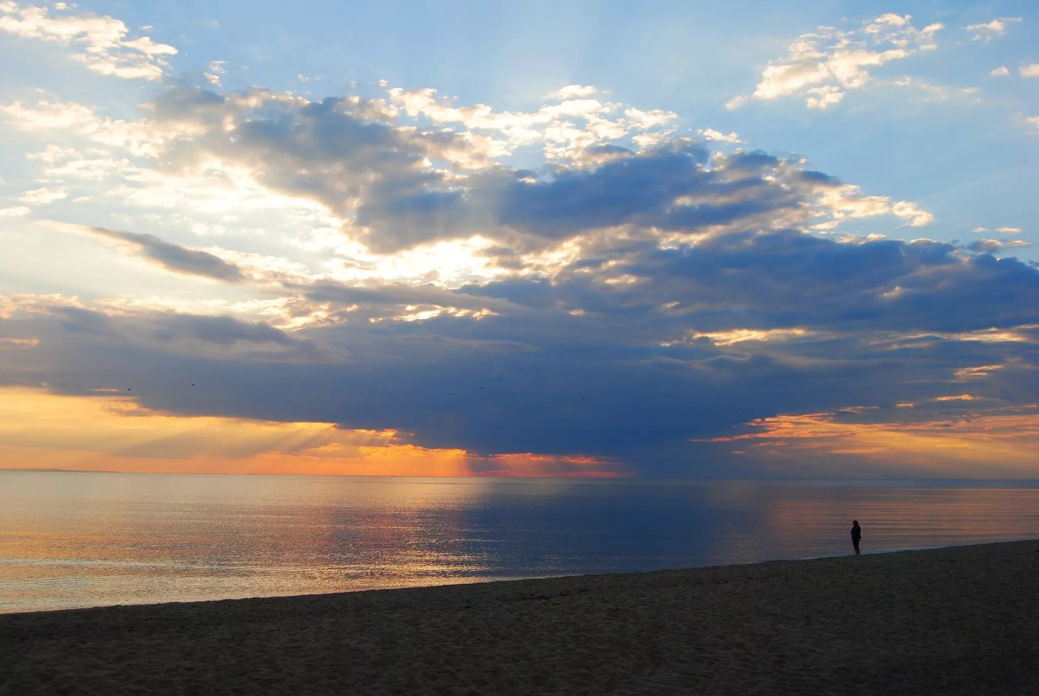 The sun setting over the sea with one person in silhouette standing on the shore.