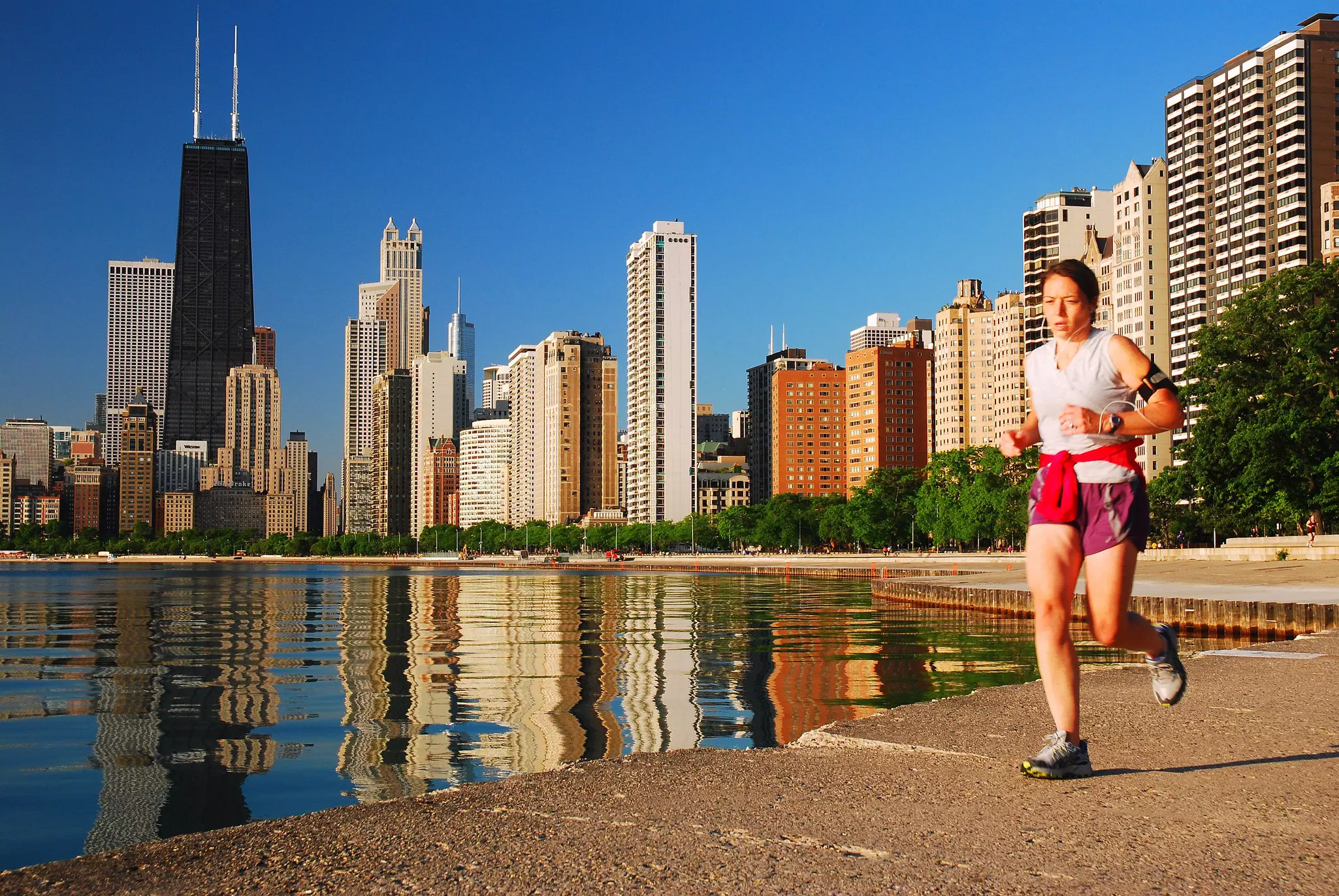 A woman jogs along a waterfront trail in a city. Tall towers line the shore behind her, and their reflections are visible in the water.