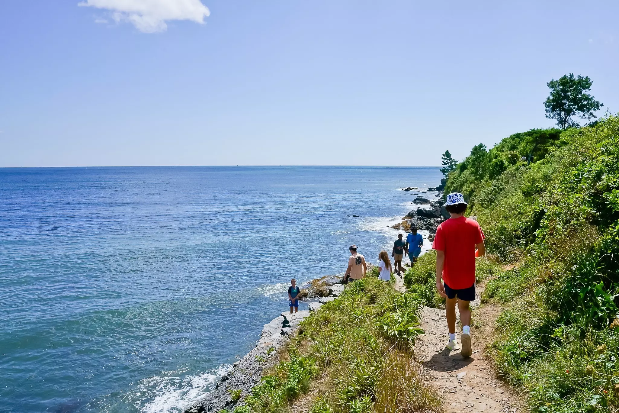People walk down a narrow dirt path to a rocky shore by the sea in Newport, Rhode Island.