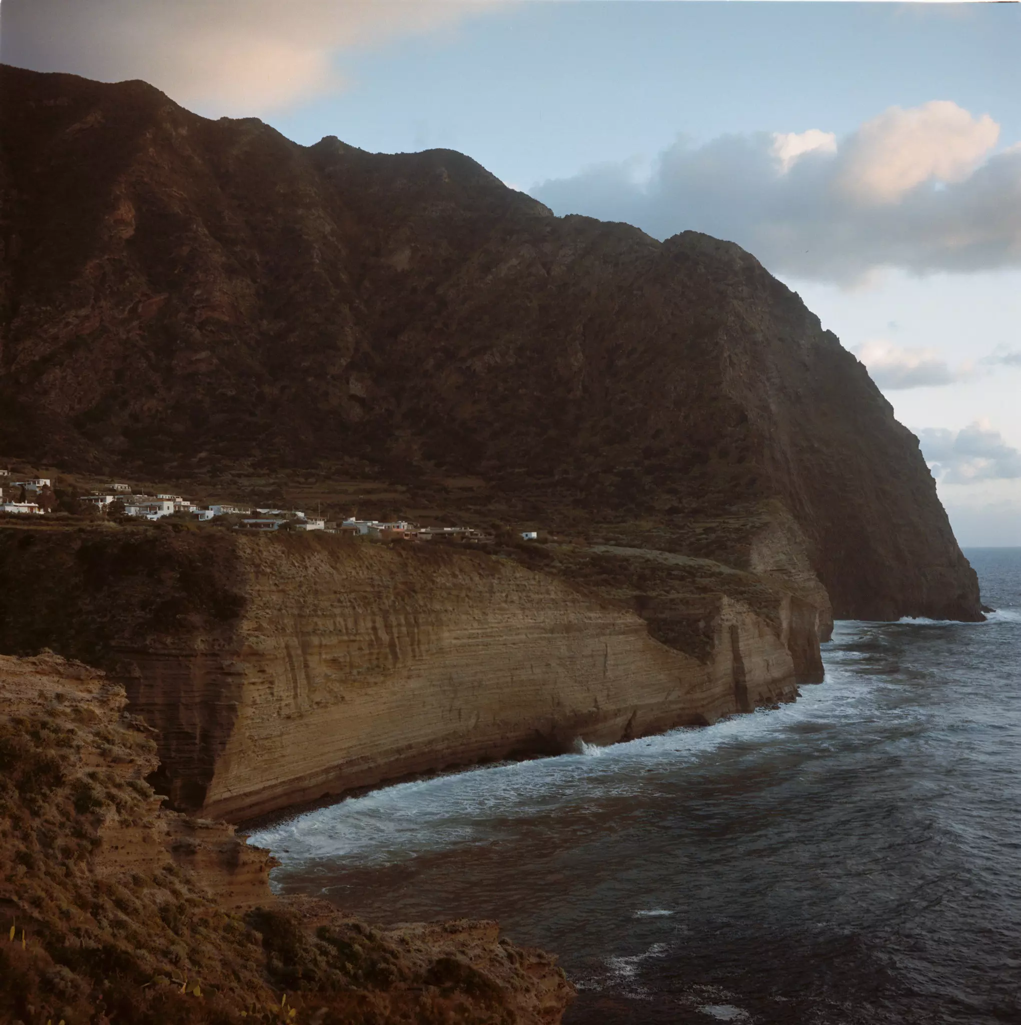 A view of a small village on a cliff over the rough sea, with a looming mountain above.