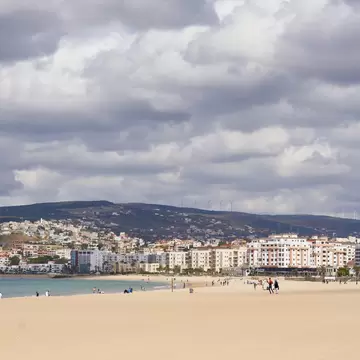 Wide sandy shore with walkers and a horse, calm turquoise water and white buildings on green hills under grey clouds