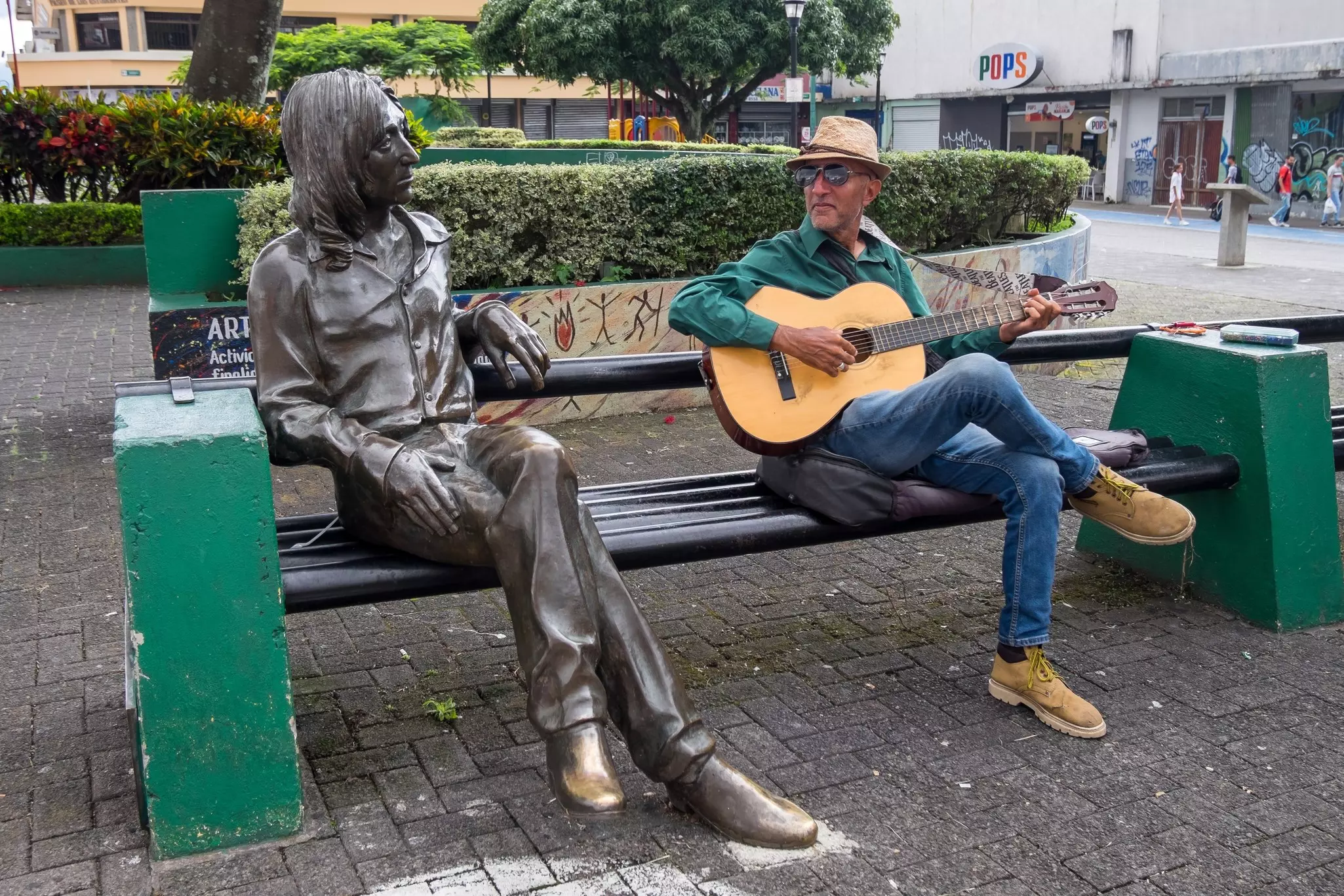 Sitting next to a life-size bronze statue, a man plays a guitar on a park bench in a city.