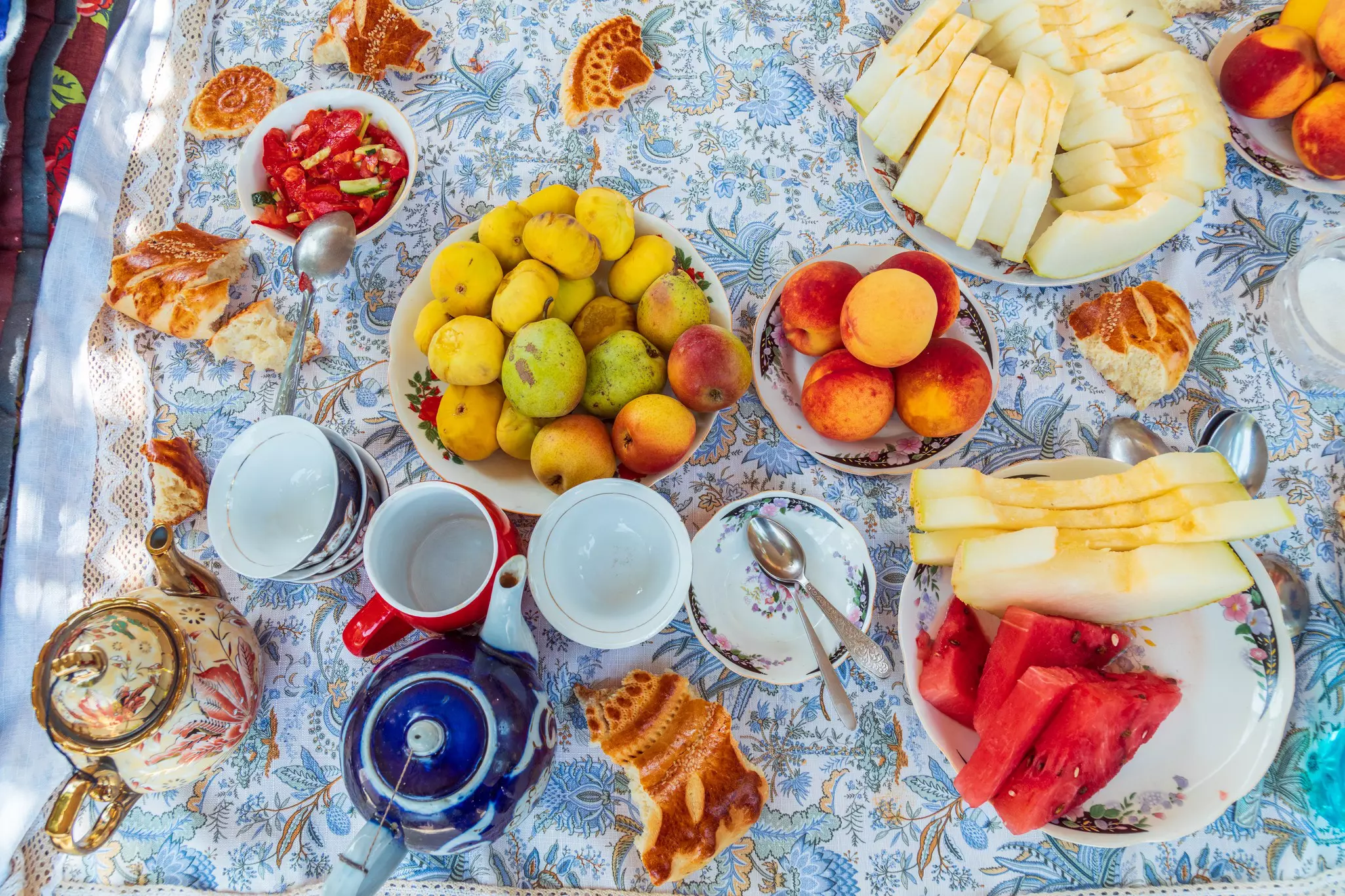 An overhead view of dishes of fruits, pastries, teacups and a teapot presented on a floral-print tablecloth