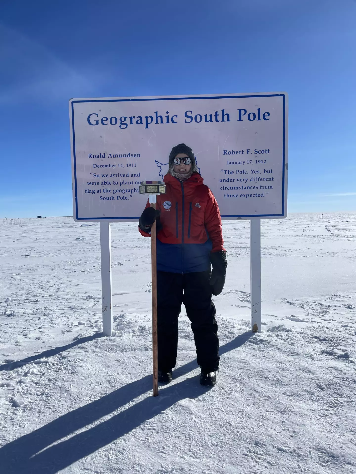 A woman stands in front of a sign in a snow field with the words "Geographic South Pole" on it.