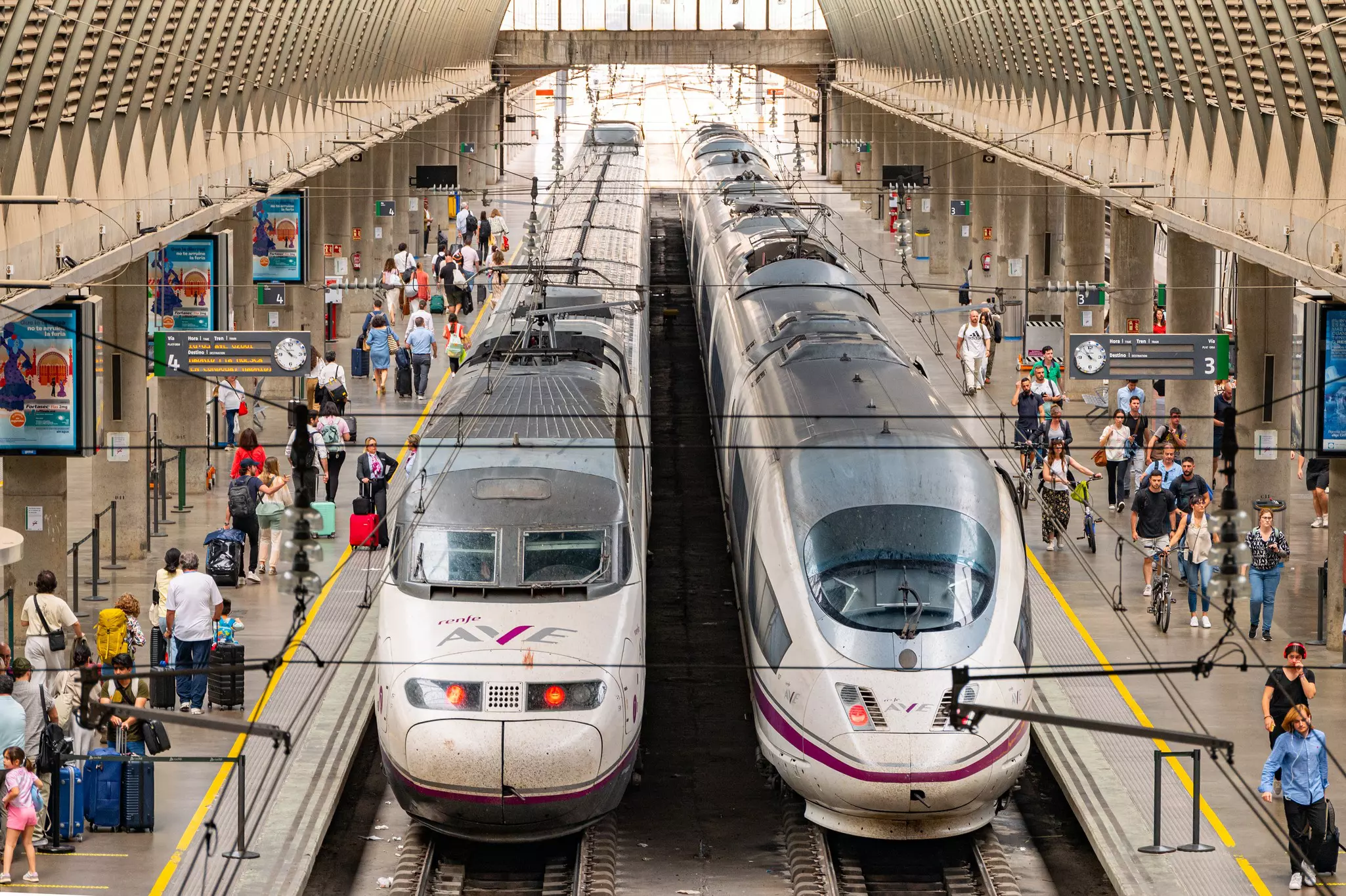 Two trains with Renfe AVE branding wait at platforms in a modern station. Passengers are boarding and disembarking.