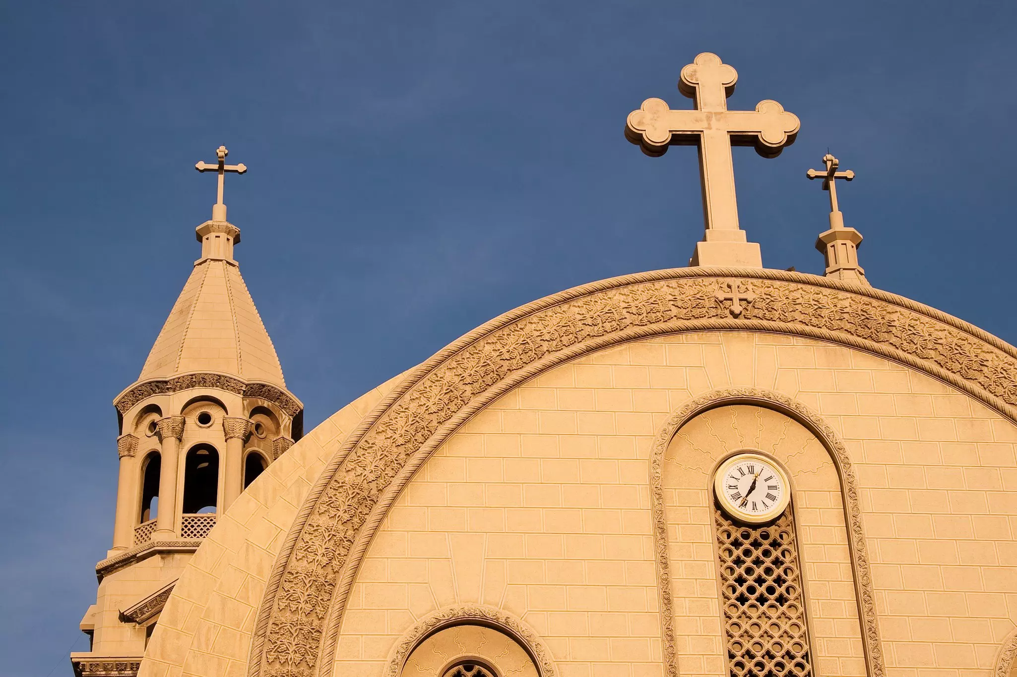 The tower, cross and clock on the facade of a tan stone church in Egypt.