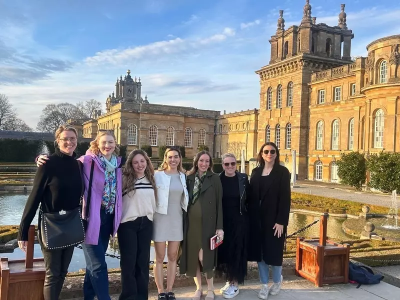 Seven people standing by a small pond in front of a large stone estate home in winter.