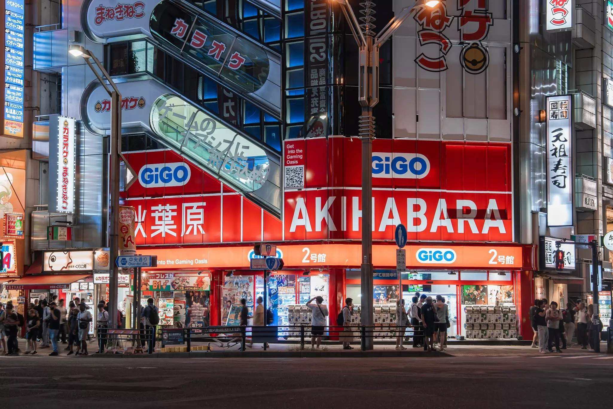 People walking on a city black in front of a glass building with lighted red signs that say "GiGO Akihabara"