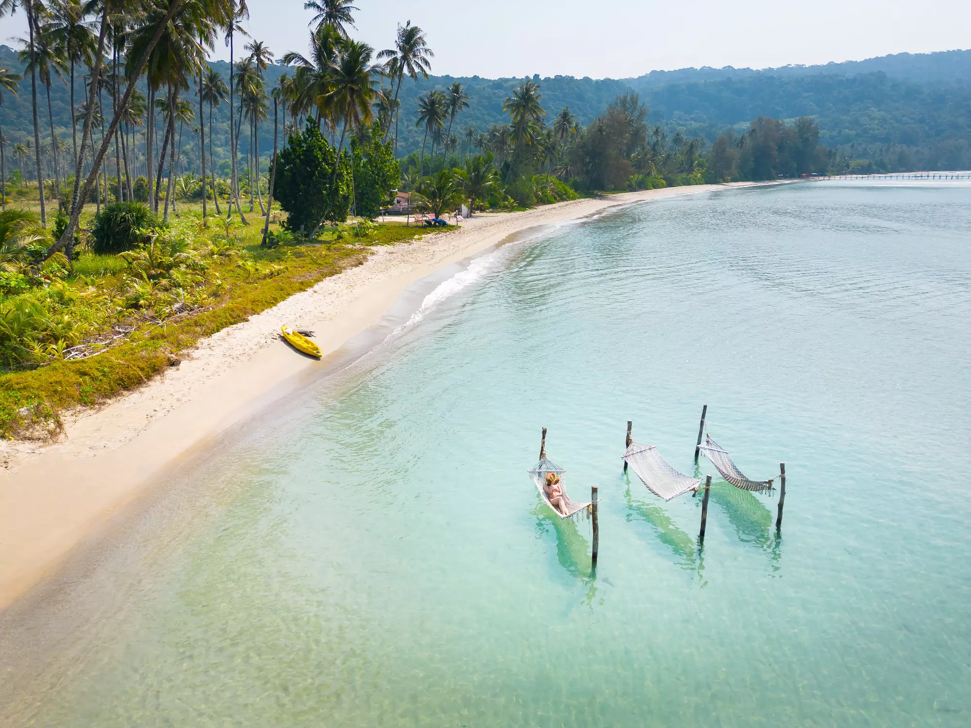 A woman lies in a hammock that's been strung on a frame in the water off an empty sandy beach backed by palm trees.