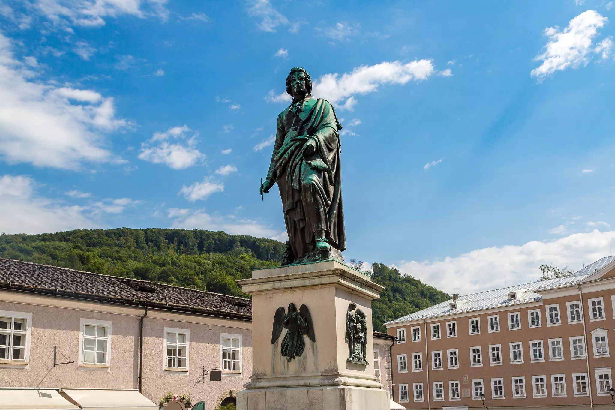 Bronze statue of Wolfgang Amadeus Mozart with a stone base and buildings and a tree-covered hill in the distance on a sunny day.