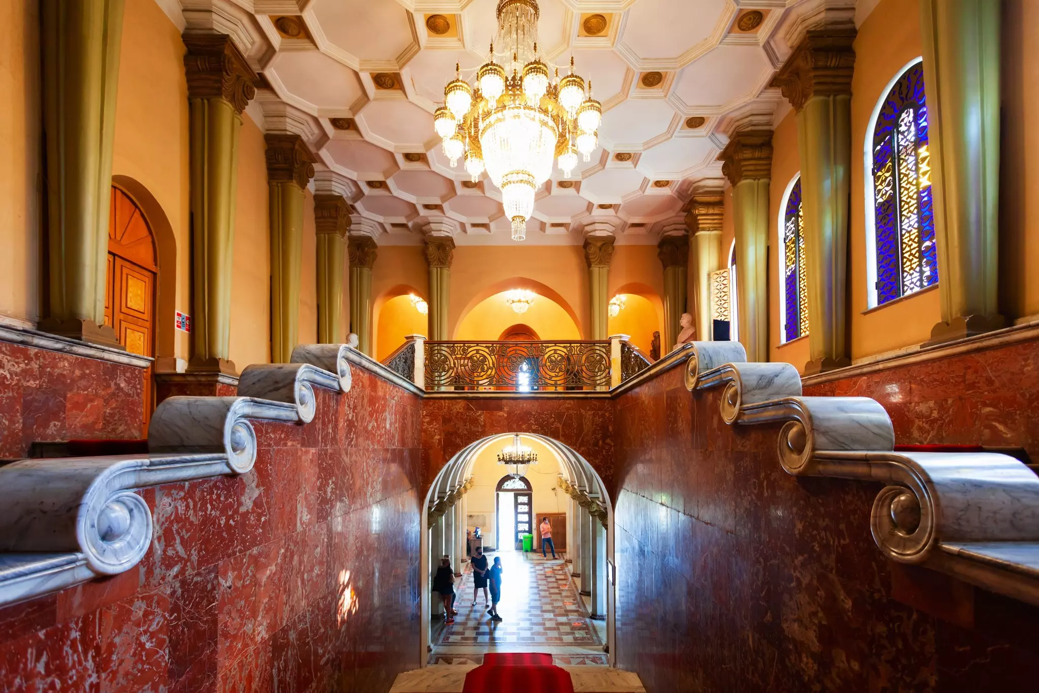 A corridor and stairwell within a museum, with ornate marble features on the bannisters, a wrought-iron balcony and large glass and gold chandelier hanging from the ceiling with hexagonol patterns.