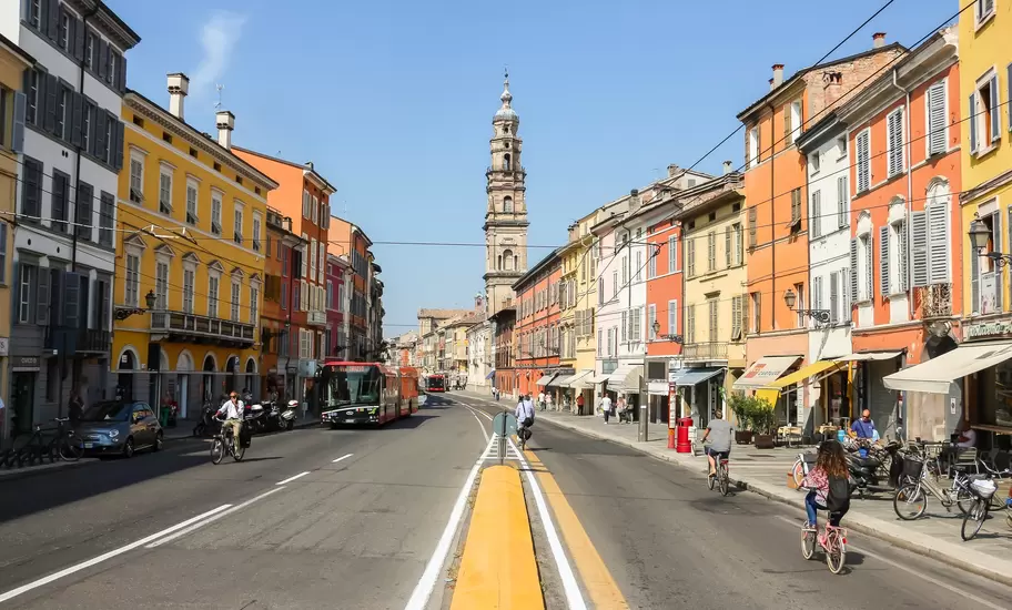 Cyclists ride down a wide street lined with pastel buildings toward a tall church bell tower in Parma, Italy