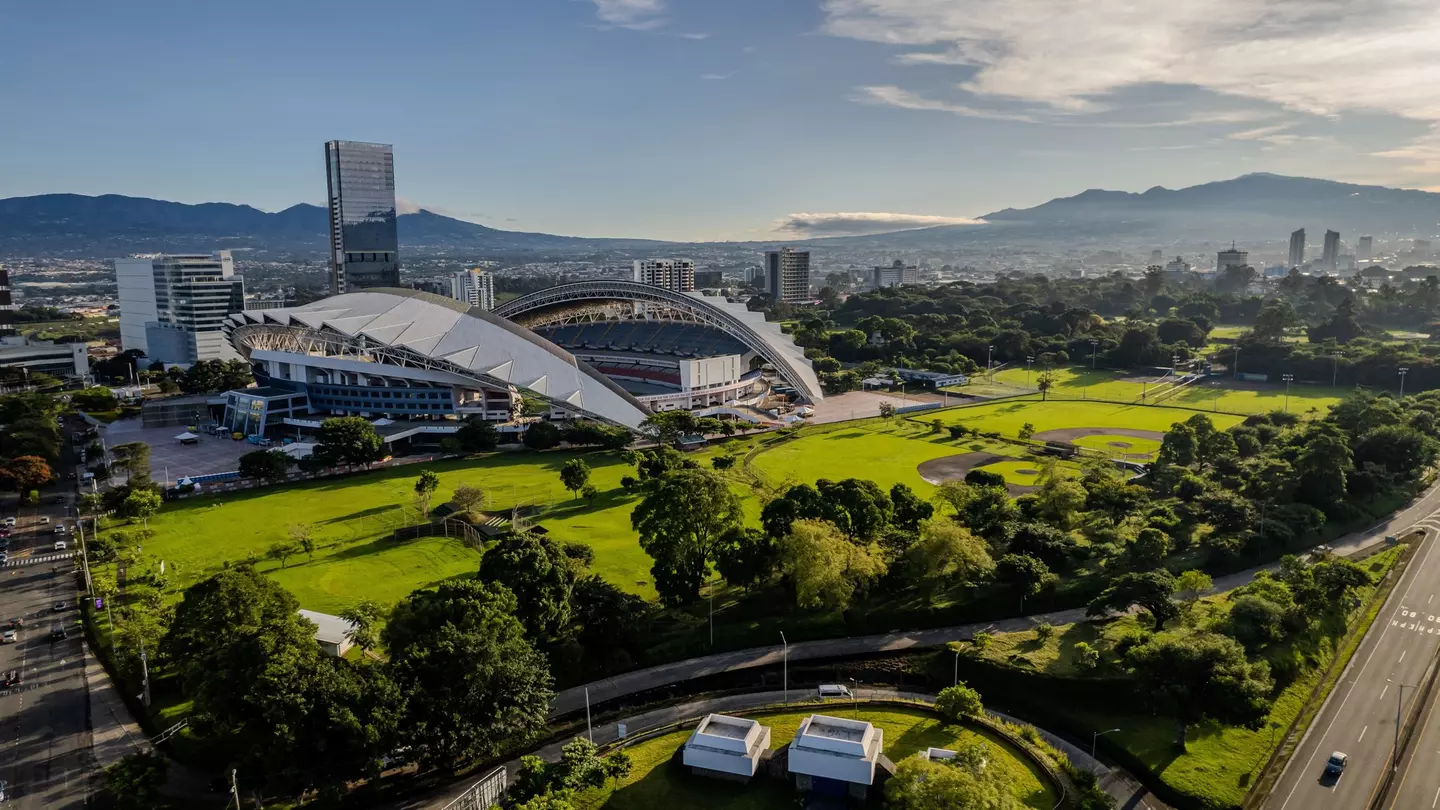 An wide shot of a green park surrounding a stadium in a busy city.