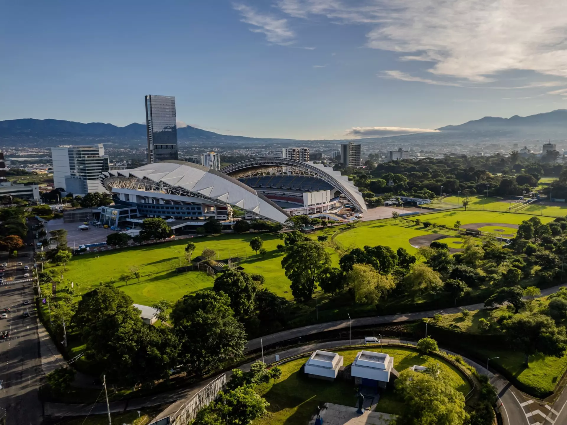 An wide shot of a green park surrounding a stadium in a busy city.