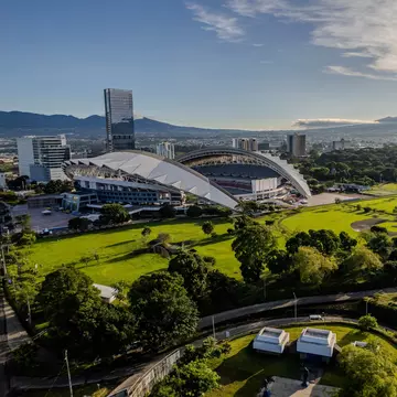 An wide shot of a green park surrounding a stadium in a busy city.