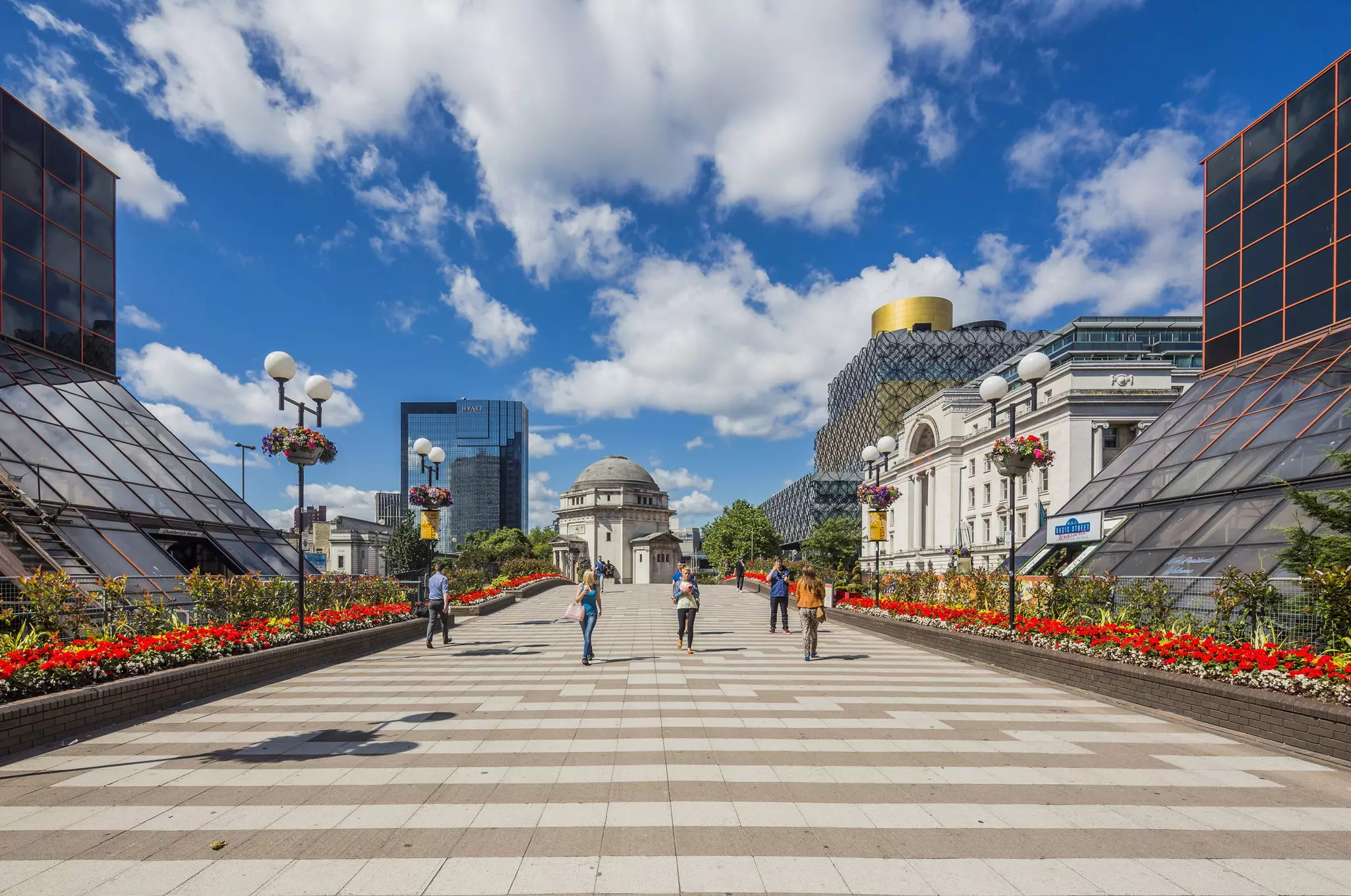 People walk along a paved path lined with flower beds in a city with a mix of old and new buildings