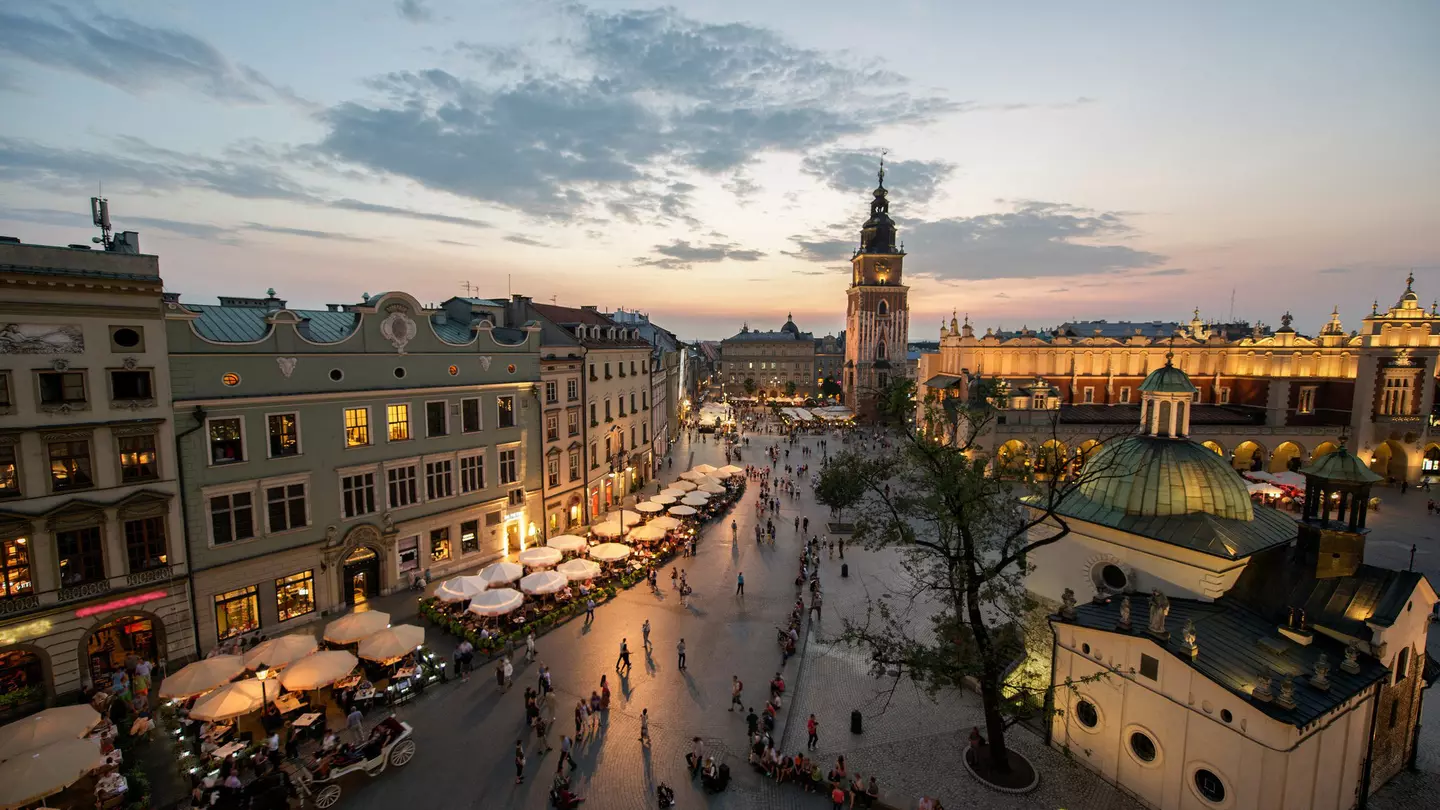 A view down over the square lined with grand buildings. The Town Hall Tower is the tallest of them all. Many outside tables at restaurants are occupied as the last light from the sun fades in the sky