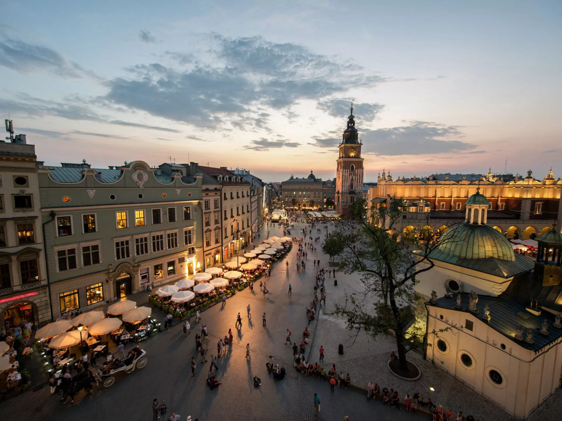 A view down over the square lined with grand buildings. The Town Hall Tower is the tallest of them all. Many outside tables at restaurants are occupied as the last light from the sun fades in the sky