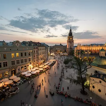 A view down over the square lined with grand buildings. The Town Hall Tower is the tallest of them all. Many outside tables at restaurants are occupied as the last light from the sun fades in the sky