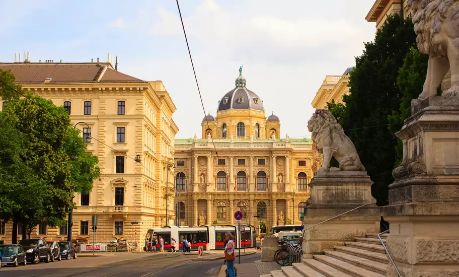 Domed Naturhistorisches Museum in Vienna seen down a street with a red-and-white tram, framed by stone lion statues on steps