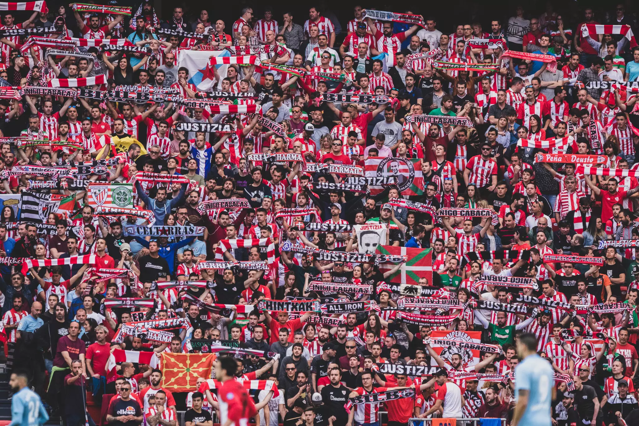 Thousands of sports fans are seen in the bleachers of a soccer stadium. They wear team colors and hold up flags and scarves.