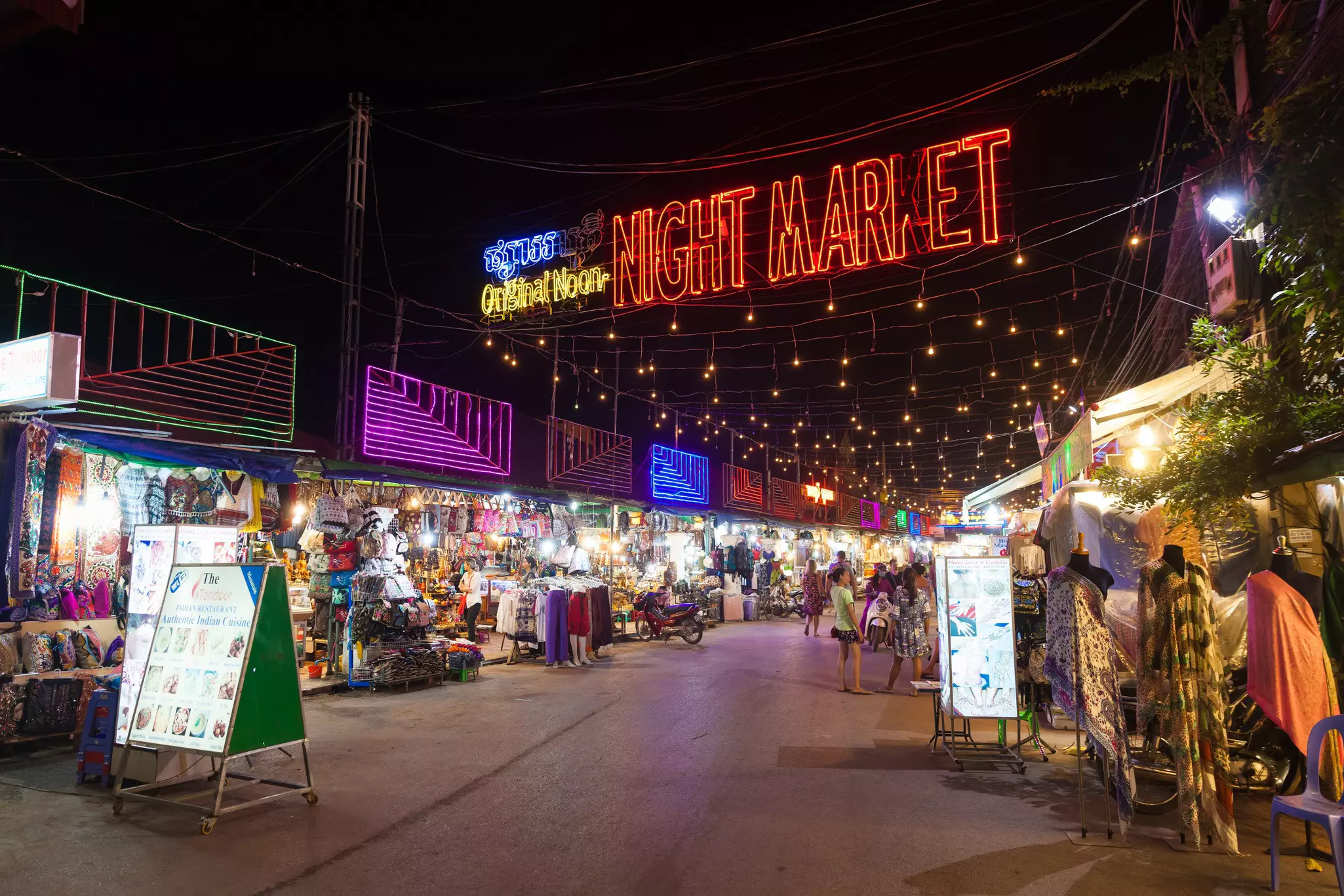 A neon sign reading "Night Market" hangs over a street with stands open at night in Siem Reap, Cambodia.