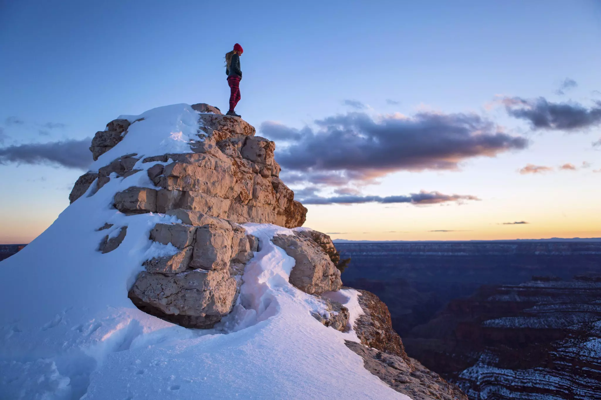 A fit, female hiker stands atop a snow-covered rocky high point while bathed in the colors of sunset at the North Rim of the Grand Canyon.