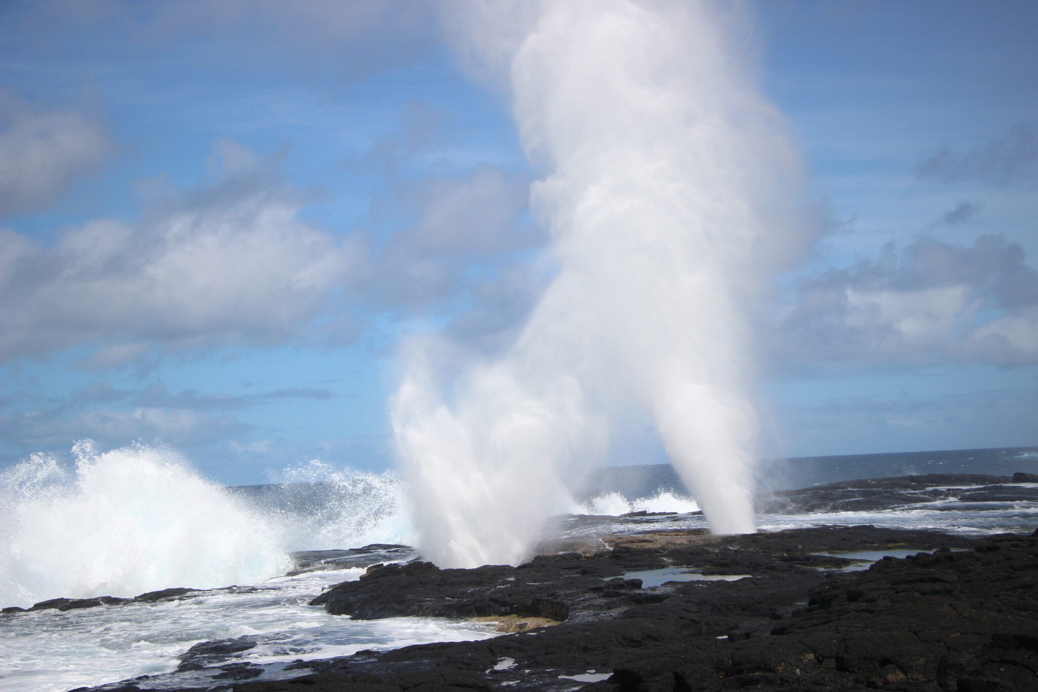 Two tall jets of white spray bursting upward from holes in a flat black lava shelf, surf breaking along the rock behind.