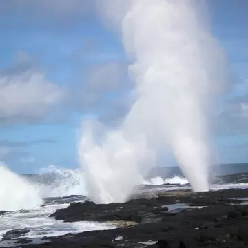 Two tall jets of white spray bursting upward from holes in a flat black lava shelf, surf breaking along the rock behind.