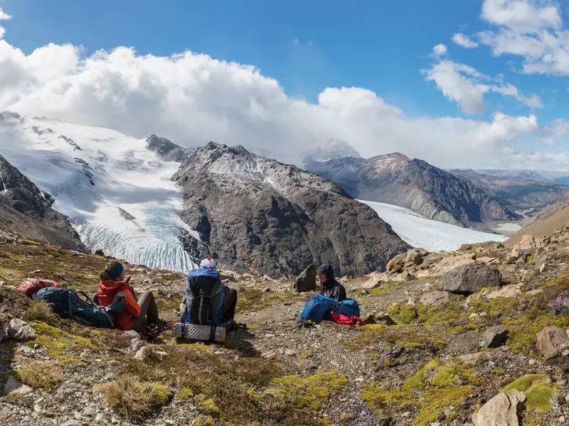 Three hikers sit on a patch of grass looking at tips of glaciers extending down a rocky mountain.