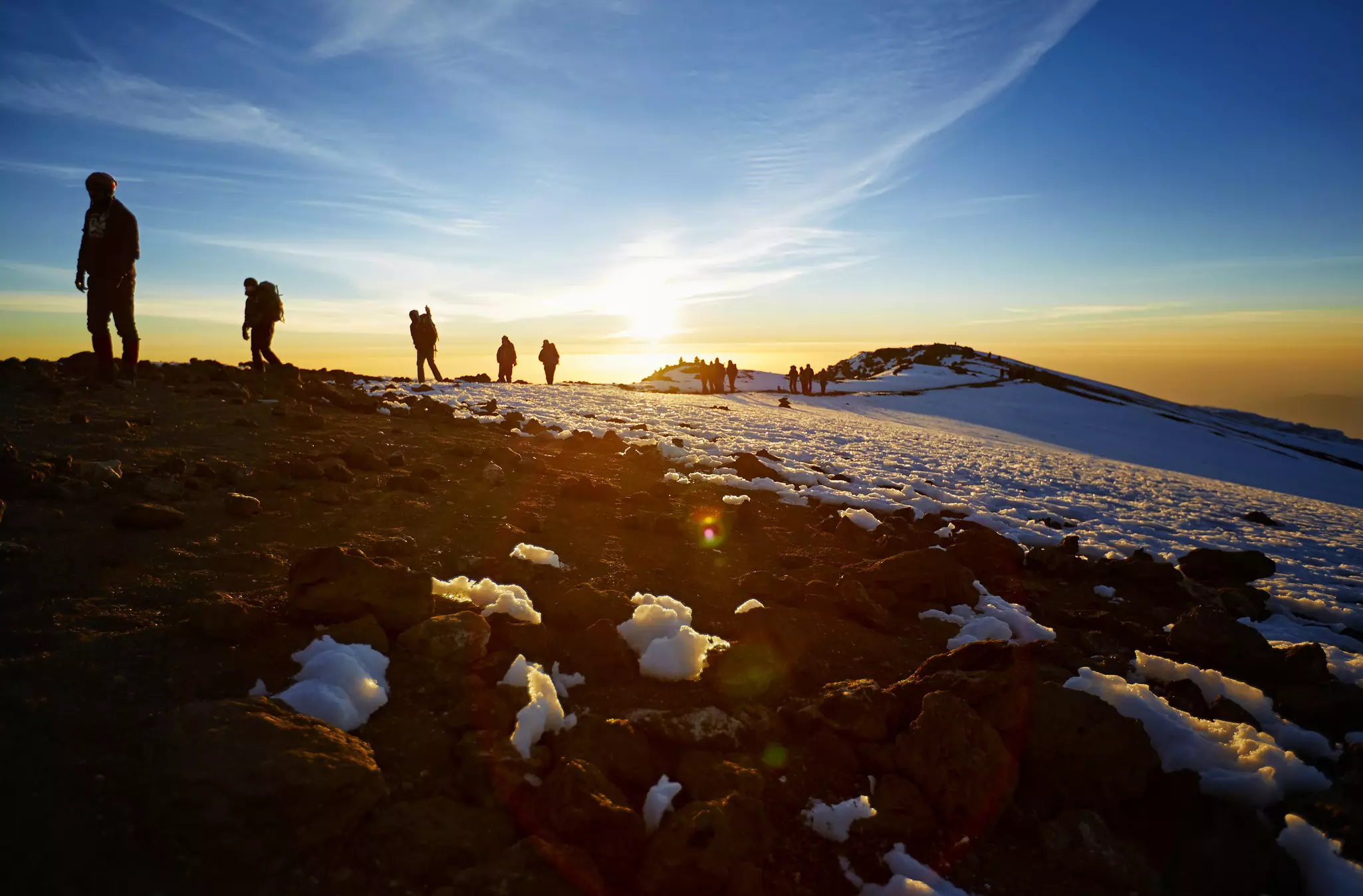 Hikers on a trail up a mountain with lumps of snow dotting the path are silhouetted in the sunrise