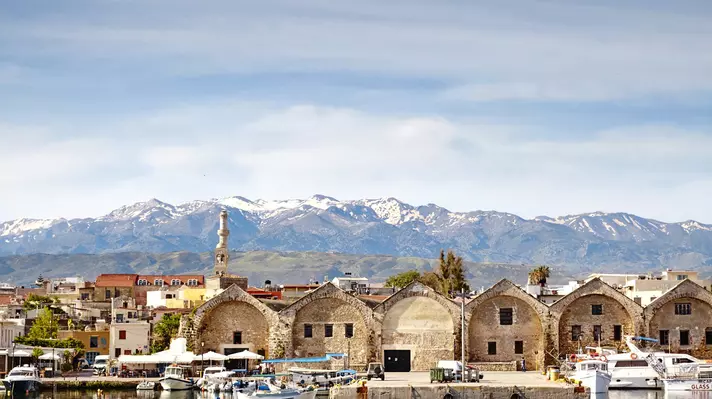 Stone buildings line a harbor in Crete with snow-capped mountains in the background. 