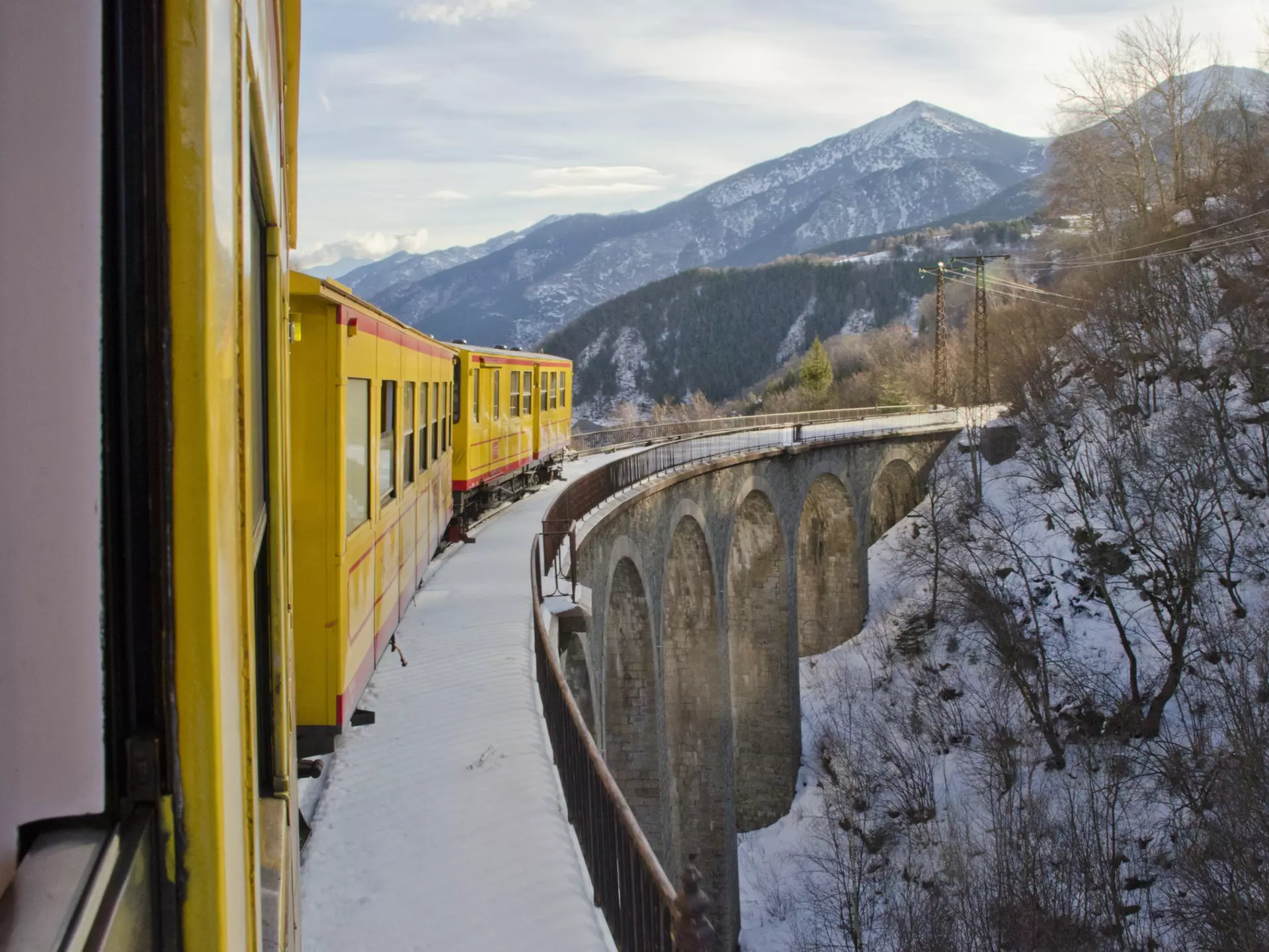 A narrow-gauge train with yellow cars navigates a viaduct through a mountain valley in winter.