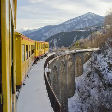 A narrow-gauge train with yellow cars navigates a viaduct through a mountain valley in winter.