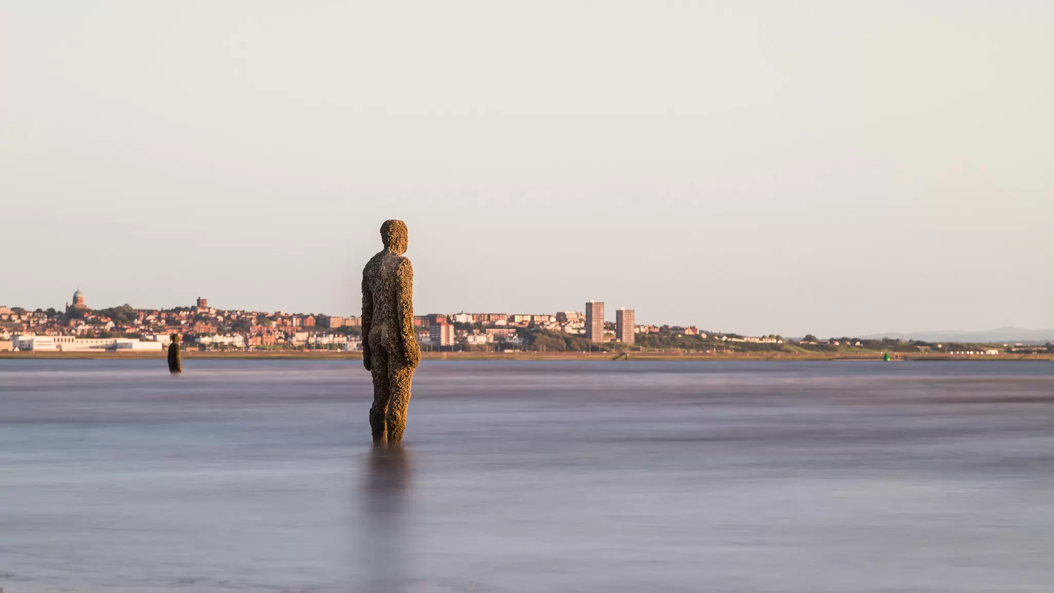 An iron statue of a full-size man stands in the water on a city beach.