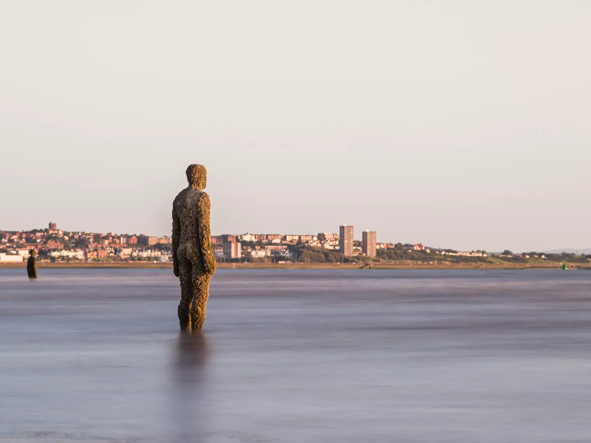 An iron statue of a full-size man stands in the water on a city beach.