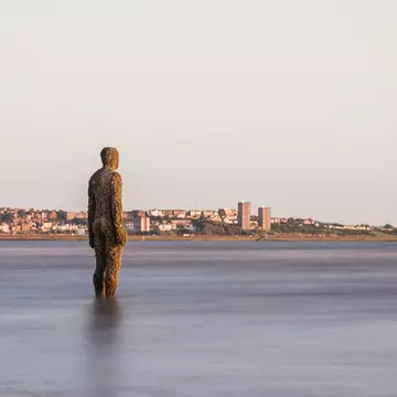 An iron statue of a full-size man stands in the water on a city beach.