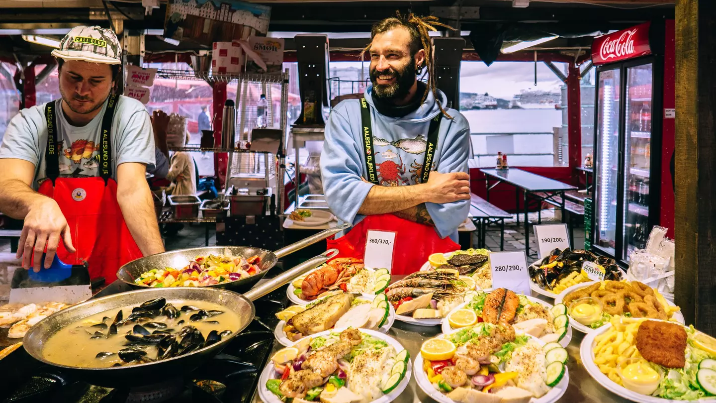 Two men wearing red aprons sell fresh fish dishes at a market in a city.