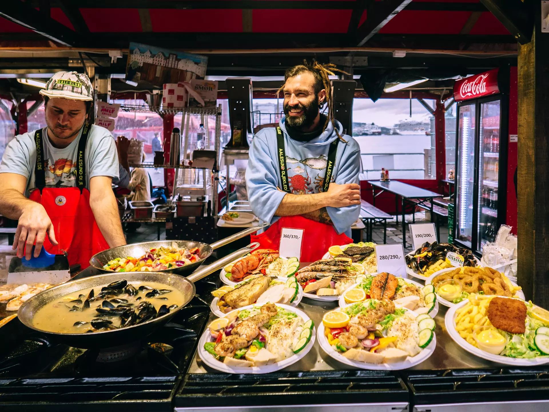Two men wearing red aprons sell fresh fish dishes at a market in a city.