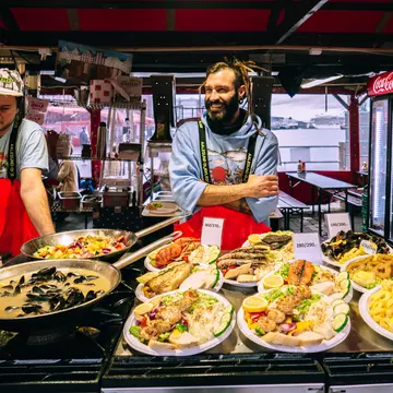 Two men wearing red aprons sell fresh fish dishes at a market in a city.