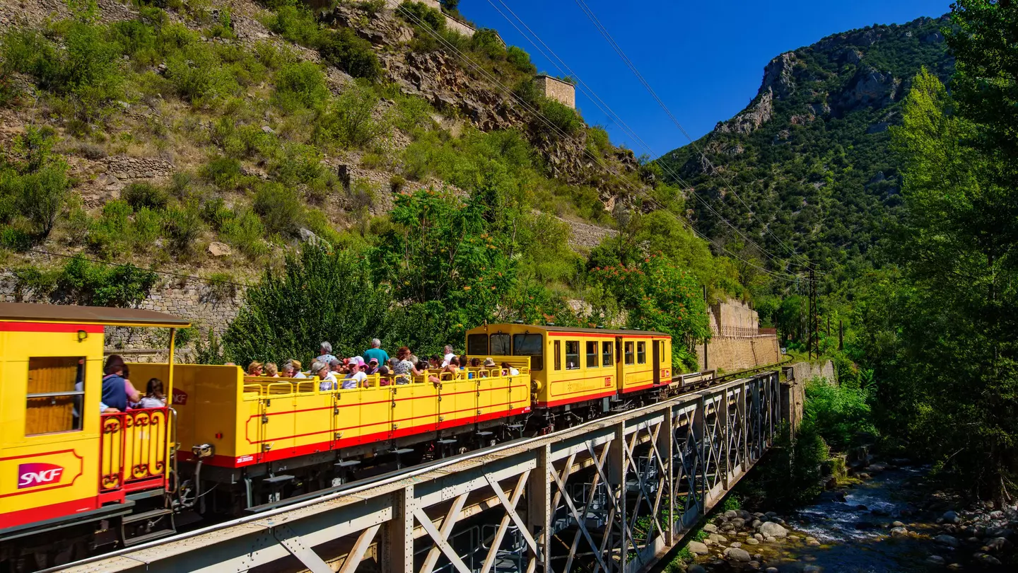 A small yellow train with passengers sat in an open-air carriage crosses a bridge over a river.