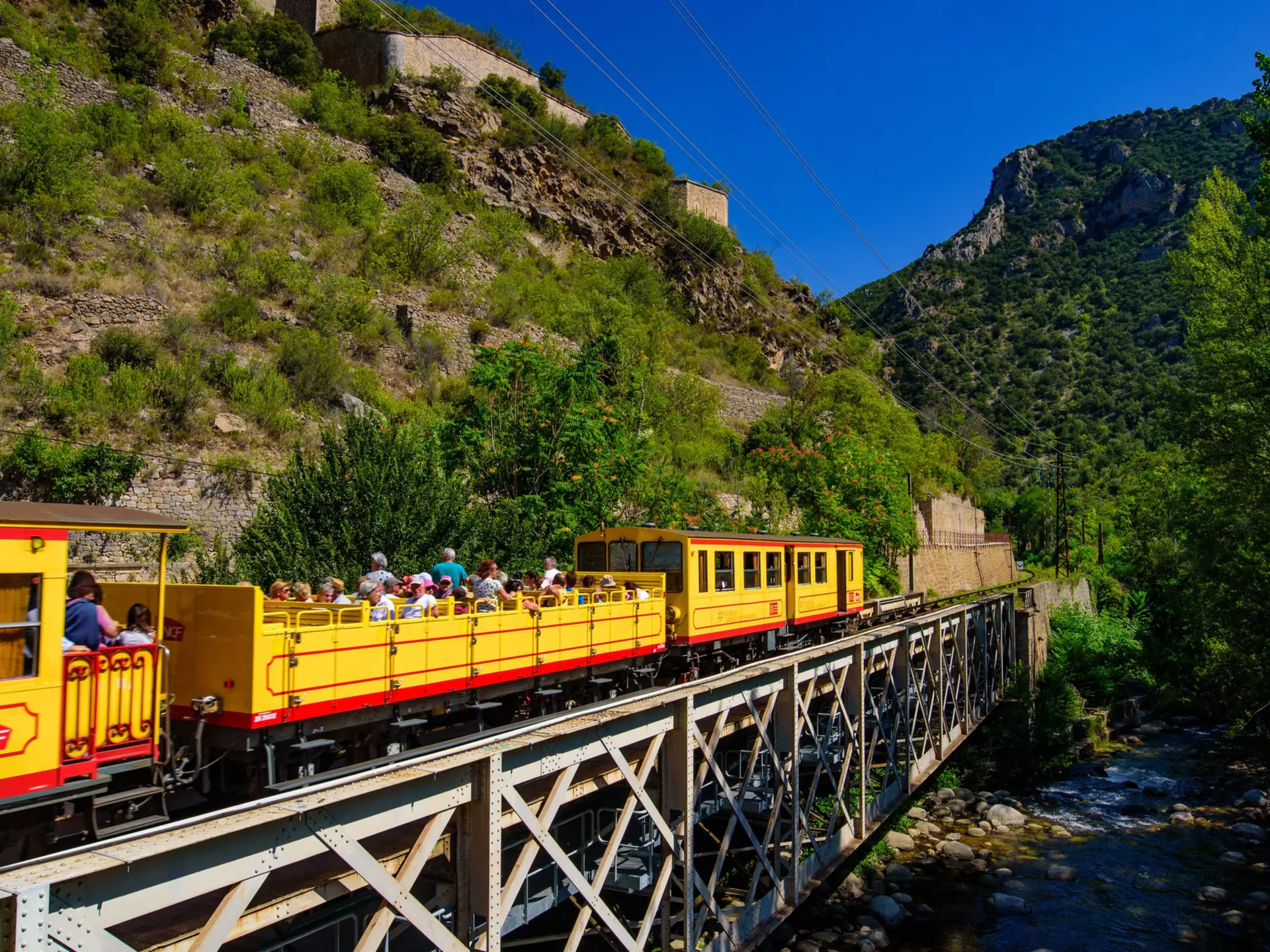 A small yellow train with passengers sat in an open-air carriage crosses a bridge over a river.
