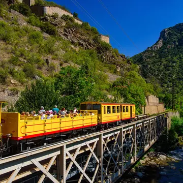 A small yellow train with passengers sat in an open-air carriage crosses a bridge over a river.