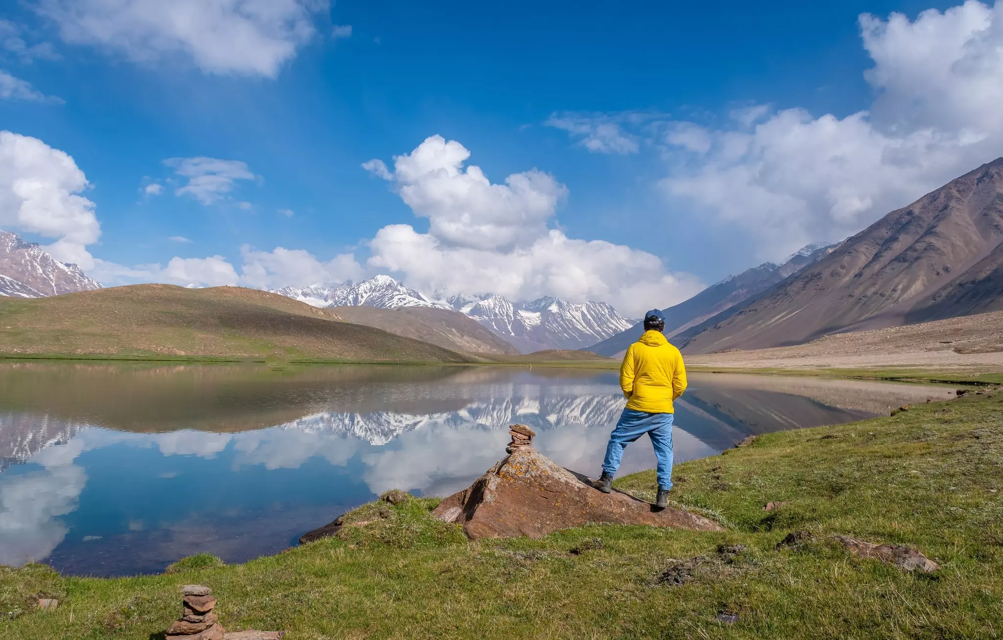 A man in a yellow windbreaker is seen from behind looking out at a still lake, the surface of which reflects snowy peaks seen in the distance.