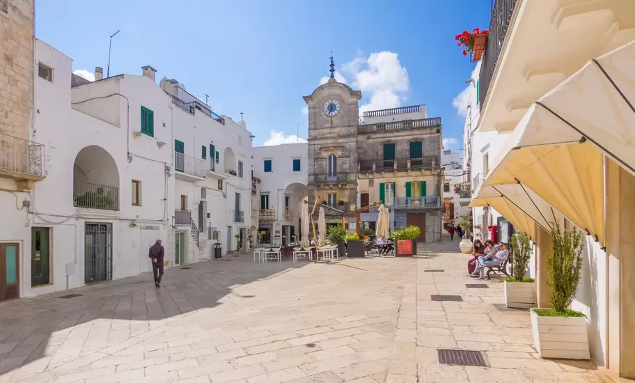 Stone clock tower above Cisternino's whitewashed main piazza, with café tables, green shutters and people on a bench