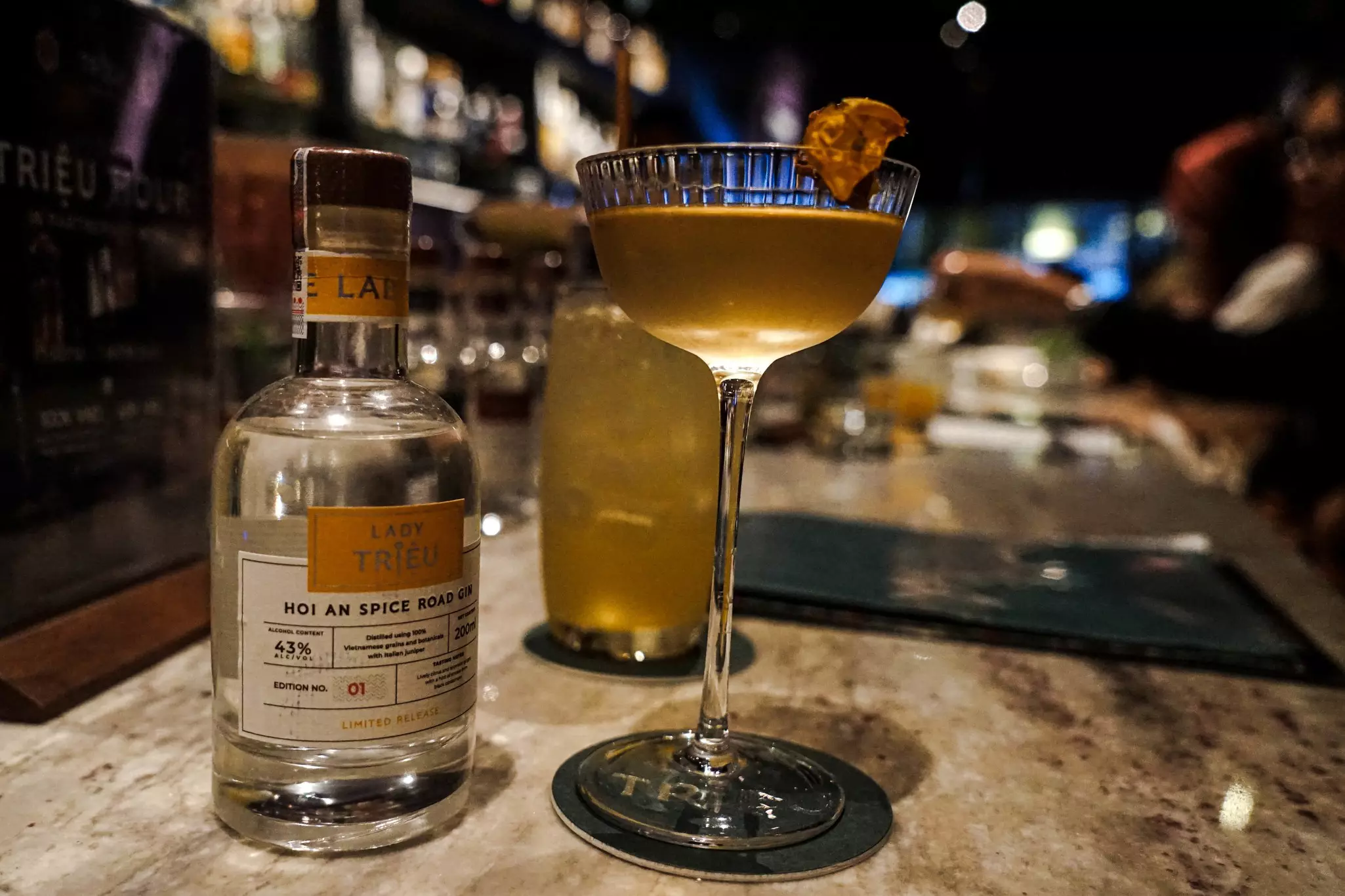 A cocktail in a stemmed glass sits on a bar next to a small bottle of gin.